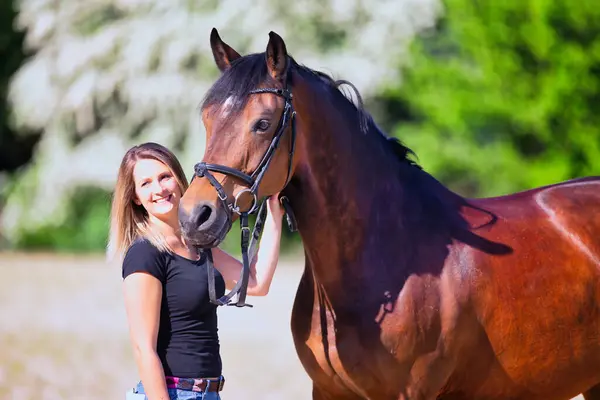 Genç bir kadın sıcak bir bahar gününün güneşinde atıyla portrede dikiliyor ve arka planda çiçek açan bir dikenli çalı var. Metin için boşluk olan manzara biçiminde resim.