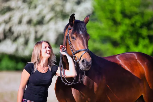 Genç bir kadın sıcak bir bahar gününün güneşinde atıyla portrede dikiliyor ve arka planda çiçek açan bir dikenli çalı var. Metin için boşluk olan manzara biçiminde resim.