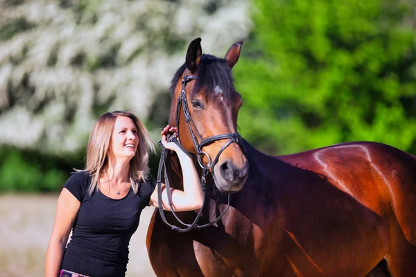 Genç bir kadın sıcak bir bahar gününün güneşinde atıyla portrede dikiliyor ve arka planda çiçek açan bir dikenli çalı var. Metin için boşluk olan manzara biçiminde resim.