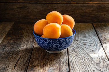 Tahta bir masanın üzerinde mavi bir seramik kasede olgun sarı kayısılar..