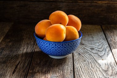 Tahta bir masanın üzerinde mavi bir seramik kasede olgun sarı kayısılar..