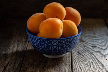 Tahta bir masanın üzerinde mavi bir seramik kasede olgun sarı kayısılar..