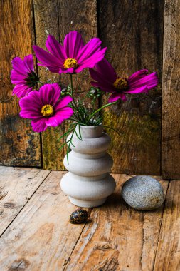 Mor çiçekli bir vazo bir kayanın yanındaki ahşap masada oturur. Çiçekler aynı yöne bakacak şekilde dizilmiş, uyum ve denge duygusu yaratıyorlar.