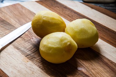 Tahta bir kesme tahtasının üzerine düzgünce yerleştirilmiş üç patates ve yanında da bir bıçak vardı. Bu çekim rahat bir mutfağın atmosferini aktarıyor, her bir malzeme,