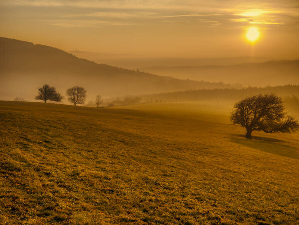 Живописный холмистый пейзаж с одинокими деревьями во время заката, вид на долину, голубое небо с высокими облаками, солнце. Осенний вечер. Биле Карпаты, Чехия.