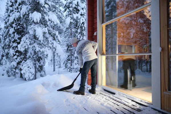 stock image mature man in hat and Scandinavian sweater clears porch of house from snow against background of snowdrifts, snow-covered trees, concept of weekend in winter forest, active lifestyle, heavy snowfall