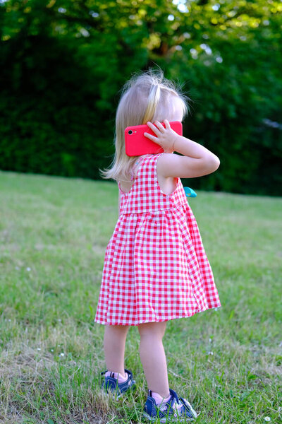 Маленький ребенок, 2-летняя девочка с белыми волосами в красном платье держит смартфон рядом с ухом, общается с друзьями, родителями, концепция детства, ранее умение обращаться с современными технологиями