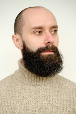 Yakın plan erkek yüzü, sakallı ve bıyıklı genç adam, bej rengi yün kazaklı 30 yıllık tatmin olmuş adam ciddi ciddi başka yere bakıyor, insan sağlığı kavramı, hayatın anlamını yansıtıyor.