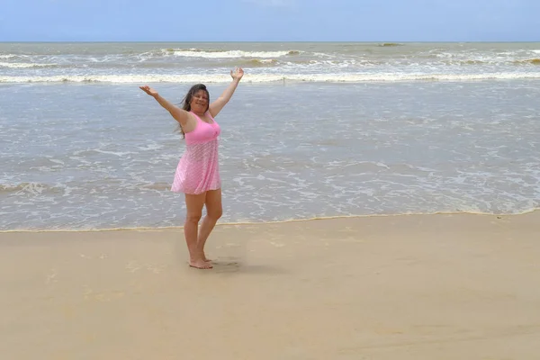 woman 50 years old with long hair in pink swimsuit stands barefoot on sandy seashore, enjoys sea breeze, takes sunbaths, blue waves, concept travel, vacation in tropical, summer sun protection