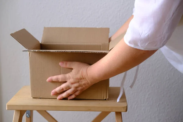 close-up of female hands open cardboard box of medium size stands on wooden stool, postal box with wrapping paper, a parcel, delivery of ordered goods, online shopping, timely delivery to customers