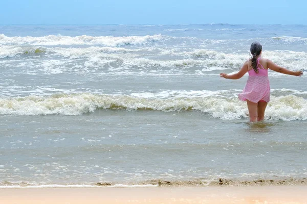 woman 50 years old with long hair in pink swimsuit with skirt enters water to swim, concept swim in waves, dangers of rip currents, safety rules in sea, travel, vacation in tropical, sun protection