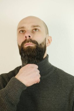 close-up male face, young bearded and mustachioed man touches beard, satisfied guy with 30 years in gray woolen sweater seriously looking away, concept of human health, reflections on meaning of life