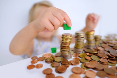 Küçük çocuk, sarışın kız 3 yaşında nakit para ile oynayan ev modeli, bozuk para, cep harçlığı, konsept ev kredisi, ev sigortası, çocukların finansal okur yazarlığı, kişisel tasarruf