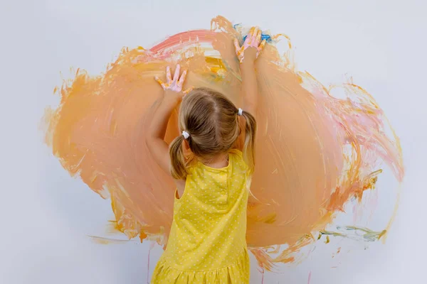 small child, blonde girl 3 years in yellow dress paints with her hands orange paint on white wall, childish naive drawing, gouache, acrylic, happiness childhood, creative development
