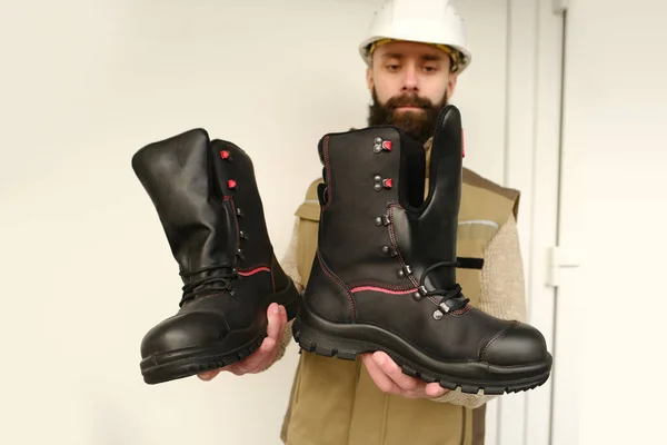 pair black Safety Shoes, work boots made of leather with reinforced cape, high top in hands of young bearded man, builder in uniform, concept of highest product quality, professional workwear