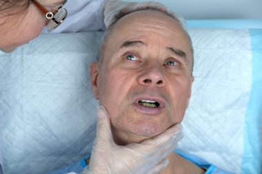 Yetişkin karizmatik bir adamın yakın çekimi, son 60 yıl mavi hastane elbisesi içinde cilt sağlığı ve güzellik kliniğinde yatıyor, doktorun elleri hastayı muayene ediyor, erkek muayenesi, yaşlanma karşıtı prosedürler