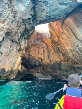 Mutlu çocuklar, turistler güneşli bir günde denizde kano kullanıyor mağaraları keşfediyor, delik kapaklı mağara, yüzme ceketli çocuk Akdeniz 'de kürek çekiyor, macera, aktif spor, harika bir tatil.