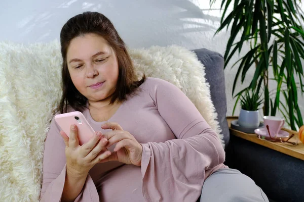 close-up of modern smartphone in female hands, middle-aged woman of 50 years old in pink sweater sits on sofa in room, reads news, communicates on Internet, concept of technology, stay at home