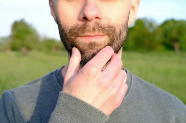 Yüzünün yakın plan kısmı, sakallı 30 yaşındaki genç adam elleriyle çenesine dokunuyor, arka planda yeşil alanlar, bahar, yaz mevsimi, sağlıklı yaşam tarzı, banliyö ve kırsal yaşam tarzı, doğa koruması