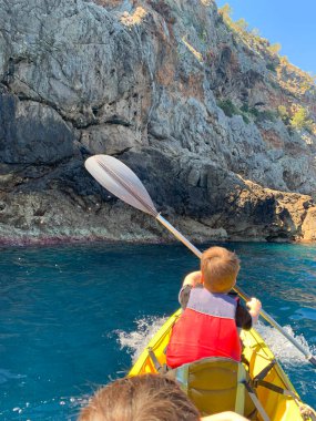Mutlu çocuk, güneşli bir günde denizde kano süren turist, 9-10 yaşlarında kova şapkalı ve yüzme ceketli sağlıklı bir çocuk Akdeniz 'de kürek çekiyor, deniz macerası, aktif spor, harika bir tatil.
