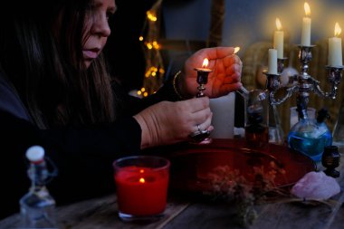 female witch holds candle in candlestick in her hands, conjures in dark room, magic items on table, concept of Halloween party, alternative medicine, esoteric practices, seance in salon of medium