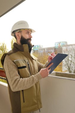 İnşaat alanındaki teftiş sırasında miğfer takan profesyonel erkek ustabaşı, üniformalı sakallı adam, müteahhit, inşaat alanında çalışan mimar not panosunda yazan, proje performansını denetleyen
