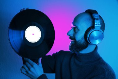 Kulaklıklı sakallı genç adamın elinde vinil disk, neon mor ve yeşil ışığa derin derin bakıyor, neon ışıklı tefekkür, kıpkırmızı parlak arka ışıklandırma