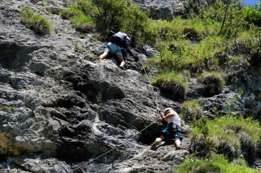 two men in body harnesses feature life-safety, attaches hooks to iron brackets, climb rock, active lifestyle, mountaineering, via ferrata in mountains of alps, life insurance in extreme sports