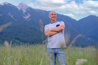 Yetişkin bir adam, şehrin yukarısındaki dağlarda 60 yaşında, arkasında gülücükler, Alpler, çayırdaki yeşil çimenler, yaşlılıktan zevk alma konsepti, seyahat, aktif yaşam tarzı, insan mutluluğu.