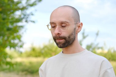 Üzgün adam, güzel altın çerçeveli gözlüklü genç karizmatik adam, bej tişörtlü, yorgun gözlüklü, arka planda yeşil bahçe, göz hastalığı konsepti, zayıf görme, doğadaki stresi azaltma.