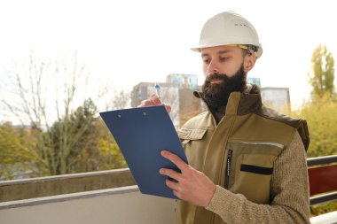 İnşaat alanındaki teftiş sırasında miğfer takan profesyonel erkek ustabaşı, üniformalı sakallı adam, müteahhit, inşaat alanında çalışan mimar not panosunda yazan, proje performansını denetleyen