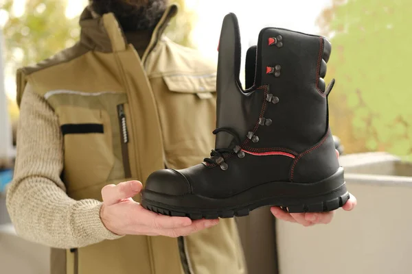 black work boots made of leather with reinforced cape, high top in hands of young bearded man, builder in uniform, concept of highest product quality, special shoes for different professions