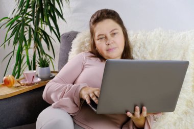 Modern dizüstü bilgisayarın kadın ellerine yakın plan çekimleri, 50 yaşında pembe kazaklı orta yaşlı bir kadın koltukta oturur, haberleri okur, internette iletişim kurar, teknoloji kavramı, evde kalır.