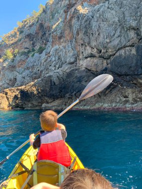 Mutlu çocuk, güneşli bir günde denizde kano süren turist, 9-10 yaşlarında kova şapkalı ve yüzme ceketli sağlıklı bir çocuk Akdeniz 'de kürek çekiyor, deniz macerası, aktif spor, harika bir tatil.