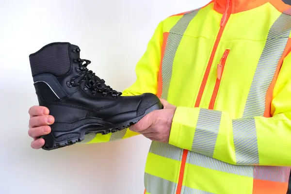 black work boots made of leather with reinforced cape, high top in hands of man, mature male mature builder, worker in yellow work clothes testing special protective professional shoes, for mountains