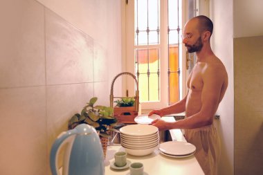 30-35 yaşlarındaki genç adam bulaşık yıkıyor, mutfak temizliği yapıyor, ev işlerine katkıda bulunuyor, ev işlerini yönetiyor, iş paylaşımı yapıyor, modern aile dinamiklerini geliştiriyor.
