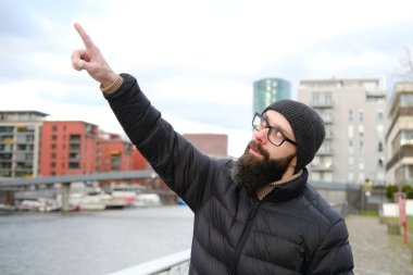 Paltolu ve şapkalı genç adam nehir kıyısında duruyor, modern yerleşim binalarına bakıyor, Frankfurt 'ta kış yürüyüşleri, Gutleitviertel bölgesinde, kıyılara demirlemiş tekne ve yatlara bakıyor.