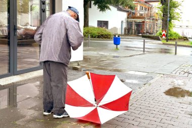 Yaşlı adam, ceketli, yağmurlu bir sokakta duruyor, cebinde para arıyor, ıslak kaldırım levhalarına kırmızı, beyaz şemsiye koyuyor, çaresiz yaşlılık kavramı.