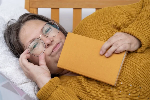 50-year-old woman in yellow sweater and glasses semi-reclined, reading boring book, Work-Life Balance, Fatigue, Feeling tired or exhausted