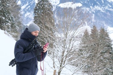 Kışlık elbiseli, fotoğraf tripodu kullanan 30 yaşında İsviçre 'deki kar örtülü dağlara dayanan profesyonel fotoğrafçılık, fotoğraf ve ekipman kavramına sahip genç bir adam.