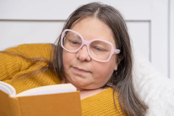 45-year-old woman in yellow sweater and glasses semi-reclined, reading Interesting book, pleasure of reading during leisure time, relaxation and enjoyment in simple activities