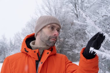 Gözlüklü genç adam, parlak turuncu ceketli, rahat bir şekilde resim parkında geziniyor, karla kaplı sakin manzaralar, kış yürüyüşü, açık hava meditasyonu, aktif yaşam tarzı, soğuk hava.