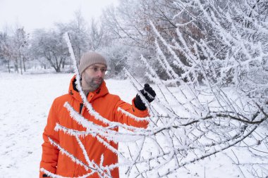 Düşünceli karizmatik bir adam. 30 yaşında, parlak turuncu ceketli, gözlüklü, doğayı izleyen, kardaki köknar ağaçları, donla kaplı ağaçlara hayran, soğuk hava, beyaz manzara, sağlıklı yaşam tarzı.