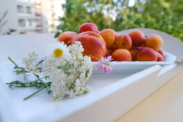 on wooden white tray plate with orange apricots, peach, bunch wild flowers on table in garden, beautiful summer still life, fruits, green trees in background, concept picnic in nature, healthy eating