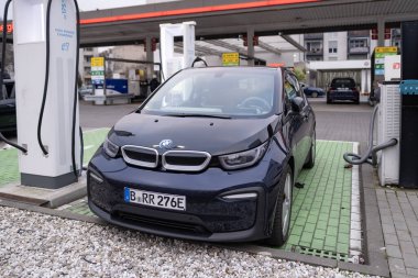 BMW i3 elektrikli araba bir elektrik şarj istasyonundan şarj ediliyor, kablo şarj ediliyor, diğer bir benzin istasyonu arka planda görünen geleneksel benzin arabaları için, Berlin, Almanya - 17 Şubat 2024