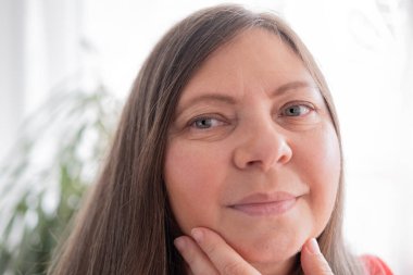 Olgun bir kadın yaşlanan yüzlere dokunur, kırışıklıklara odaklanır, yüz hatlarındaki sarkmalar ve değişimler, doğal süreçlerin yaşlanmasını, kişisel bakım uygulamalarını, rutin cilt bakımını yansıtır.