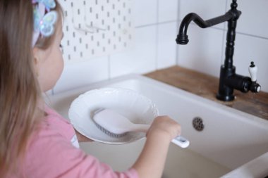 Küçük bir çocuk, beş yaşında sevimli bir kız bulaşıkları yıkıyor, tabakları mutfakta yıkıyor bulaşıkları yıkamaya odaklanıyor, erken yaştan itibaren bağımsızlığı öğretmenin önemini gösteriyor.