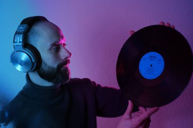Kulaklıklı sakallı genç adamın elinde vinil disk, neon mor ve yeşil ışık, DJ kültürü, neon ışıklı tefekkür, kıpkırmızı parlak arka ışıklandırma,
