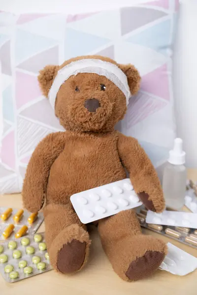 柔らかいおもちゃ 医学療法および小児科を通して散らばった薬および丸薬 ケアおよび処置の子供によって囲まれる包まれた頭部が付いているテディベア — ストック写真