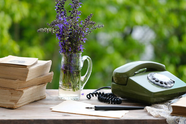 old books, rotary telephone, wild flowers on old wooden table in garden, remote work in summer in retro style, beautiful blurred natural landscape in background, being relaxed and free from stress